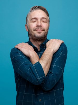 hombre-de-mediana-edad-con-cabello-gris-en-camisa-de-color-oscuro-que-parece-complacido-y-feliz-abrazandose-si-mismo-de-pie-sobre-fondo-azul