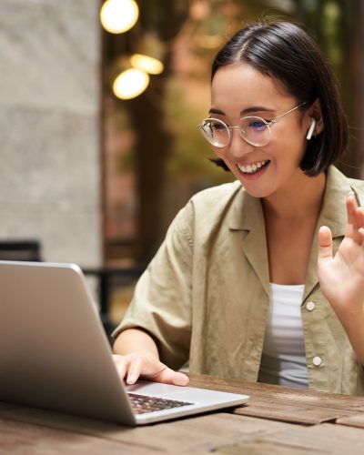 Online meeting. Young urban girl, asian woman talking with laptop, video chat, gesturing, sitting in an outdoor cafe and working remotely.